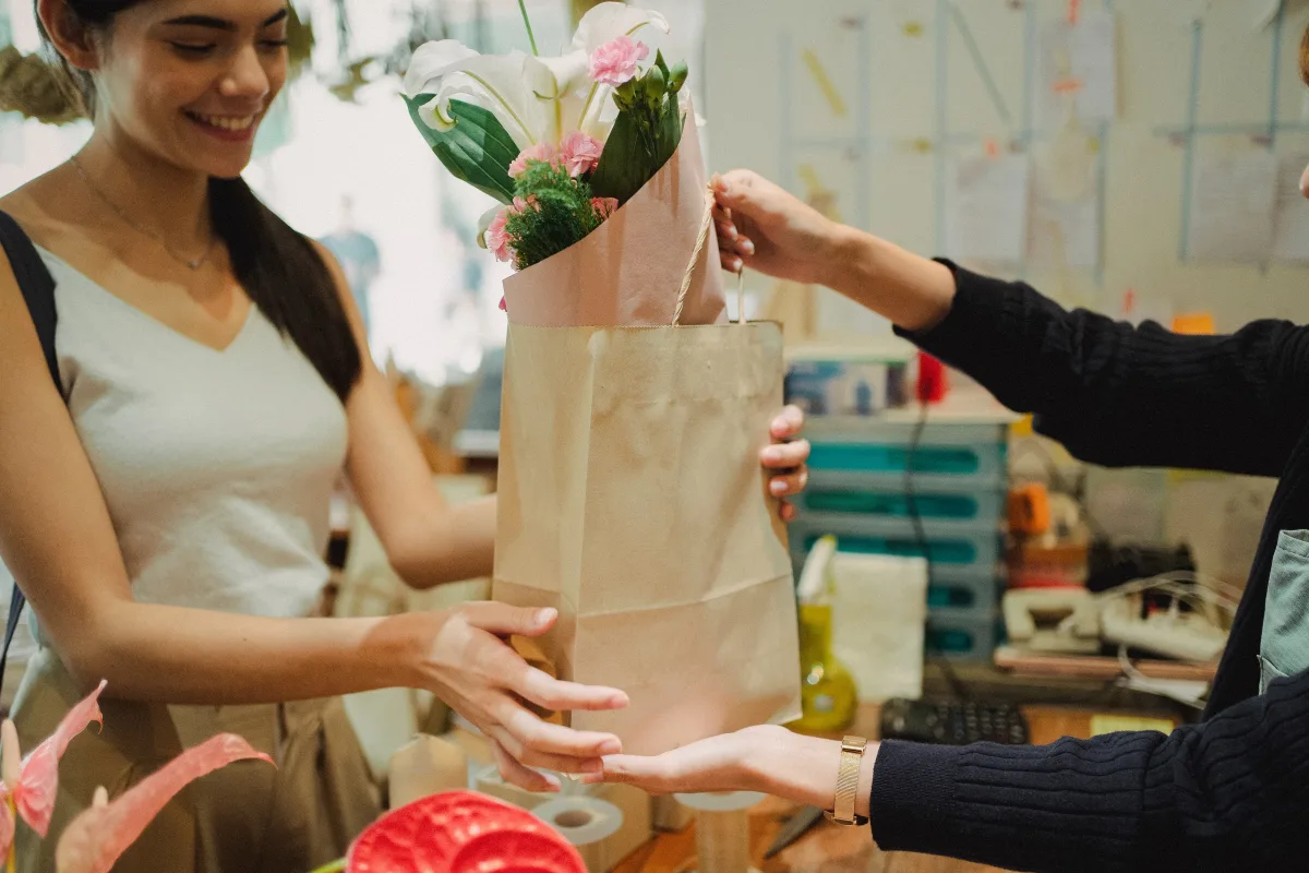 Smiling florist handing a paper bag with flowers to a customer in a bright store